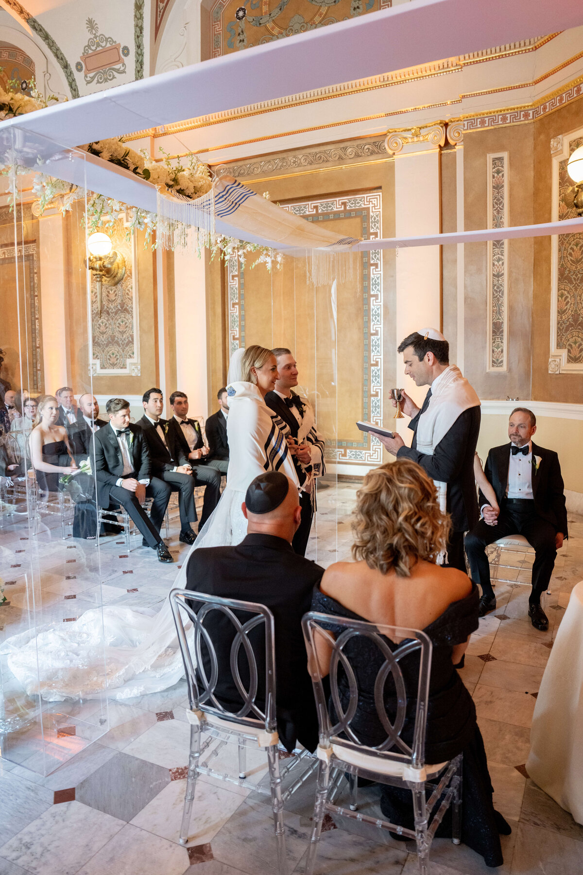 A couple stands under a chuppah, surrounded by guests seated on transparent chairs in an ornate room. The officiant reads from a book, while a man wearing a kippah sits in the foreground. The setting is elegant and formal.