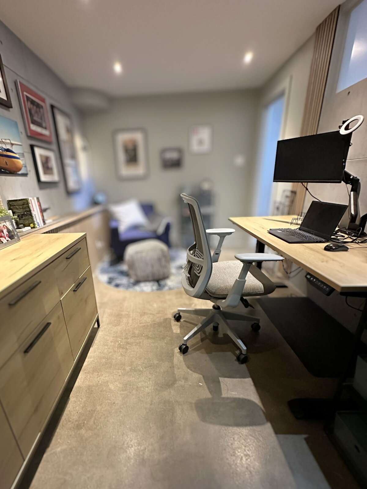 Modern home office with sit-stand desk, ergonomic chair, articulating monitor arms, wood credenza, and a cozy armchair in the background by Urbanomic Interiors & Design, Ottawa West.