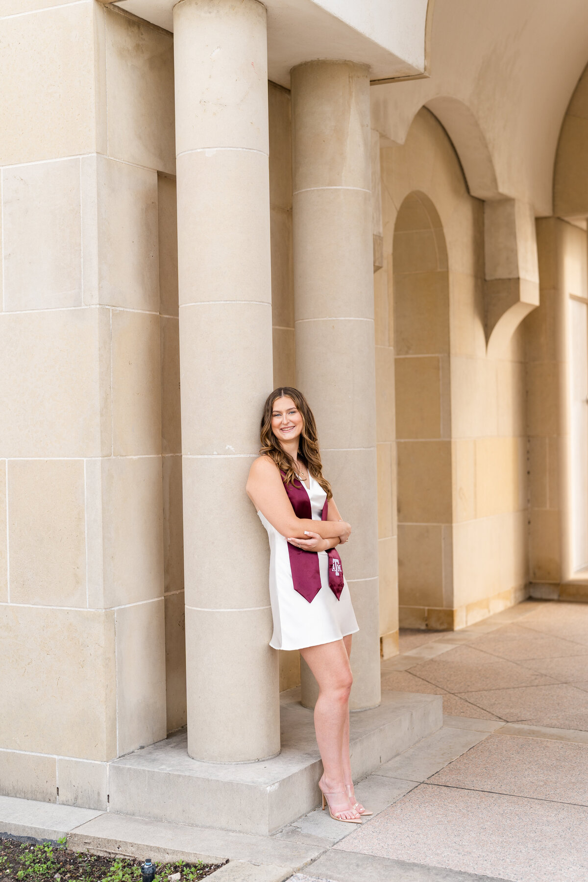Texas A&M senior girl crossing arms and leaning against column  while wearing white dress and Aggie stole and smiling away at Bell Tower