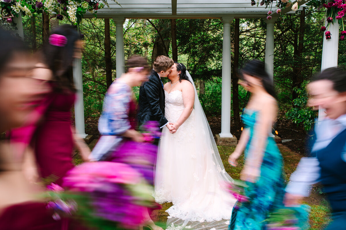Two brides kissing while bridal party passing by in a blur