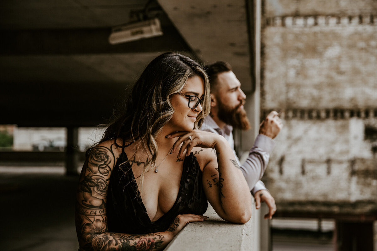 Parking garage engagement session in downtown London, ON. The man and woman are casually looking over the edge with a little distance between them. The man is smoking a cigarette looking in the distance. The woman is resting her chin on her hand and casually looking down below.