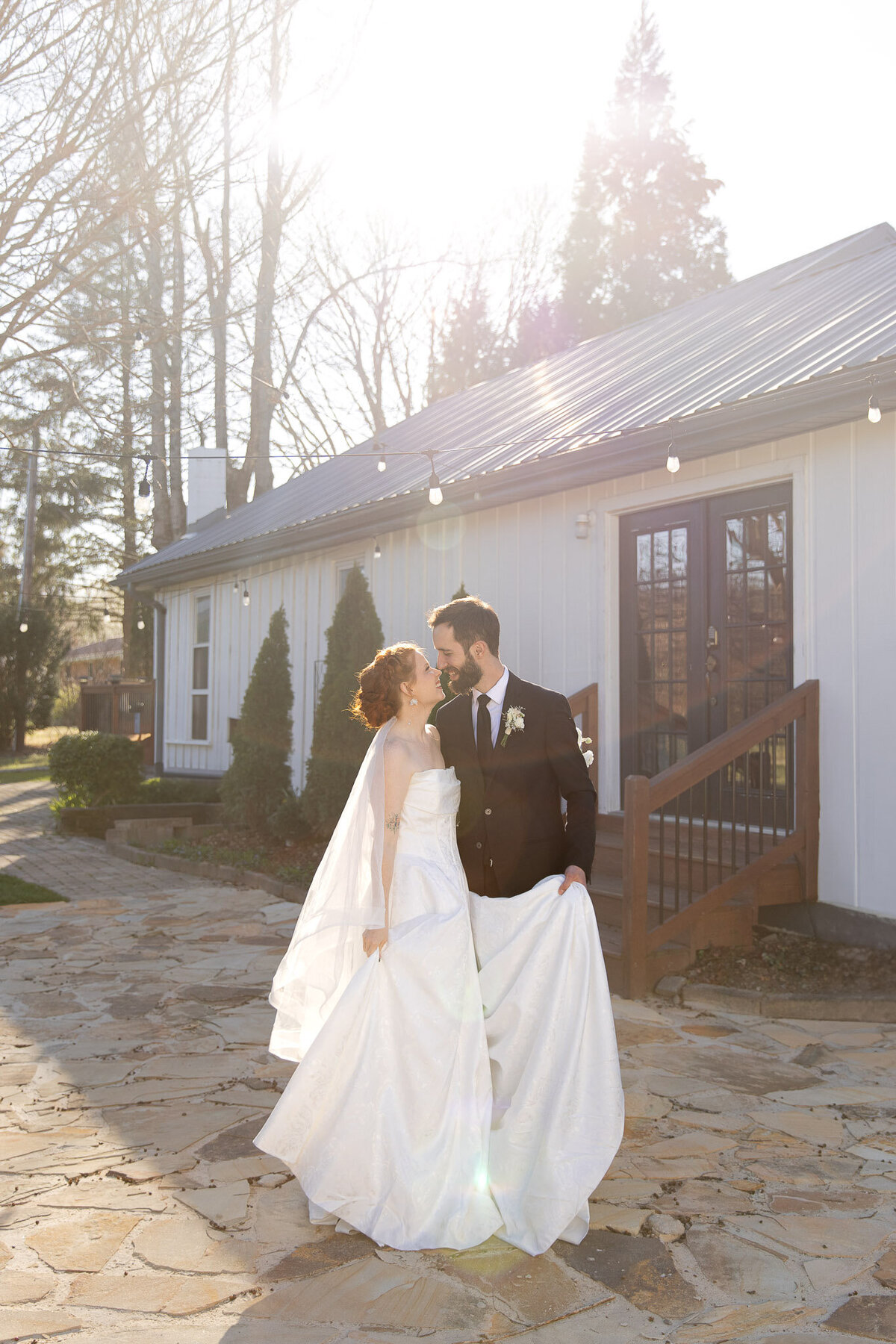 Holding his bride's dress in one hand, a groom holds the waist of his bride in the other as they romantically look into each other's eyes.