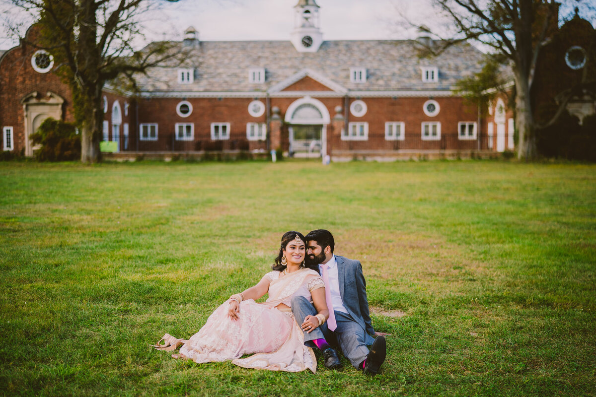 Engagement couple sitting on the grass in indian clothing in front of the lloyd harbor equestrian center at caumsett state park in long island