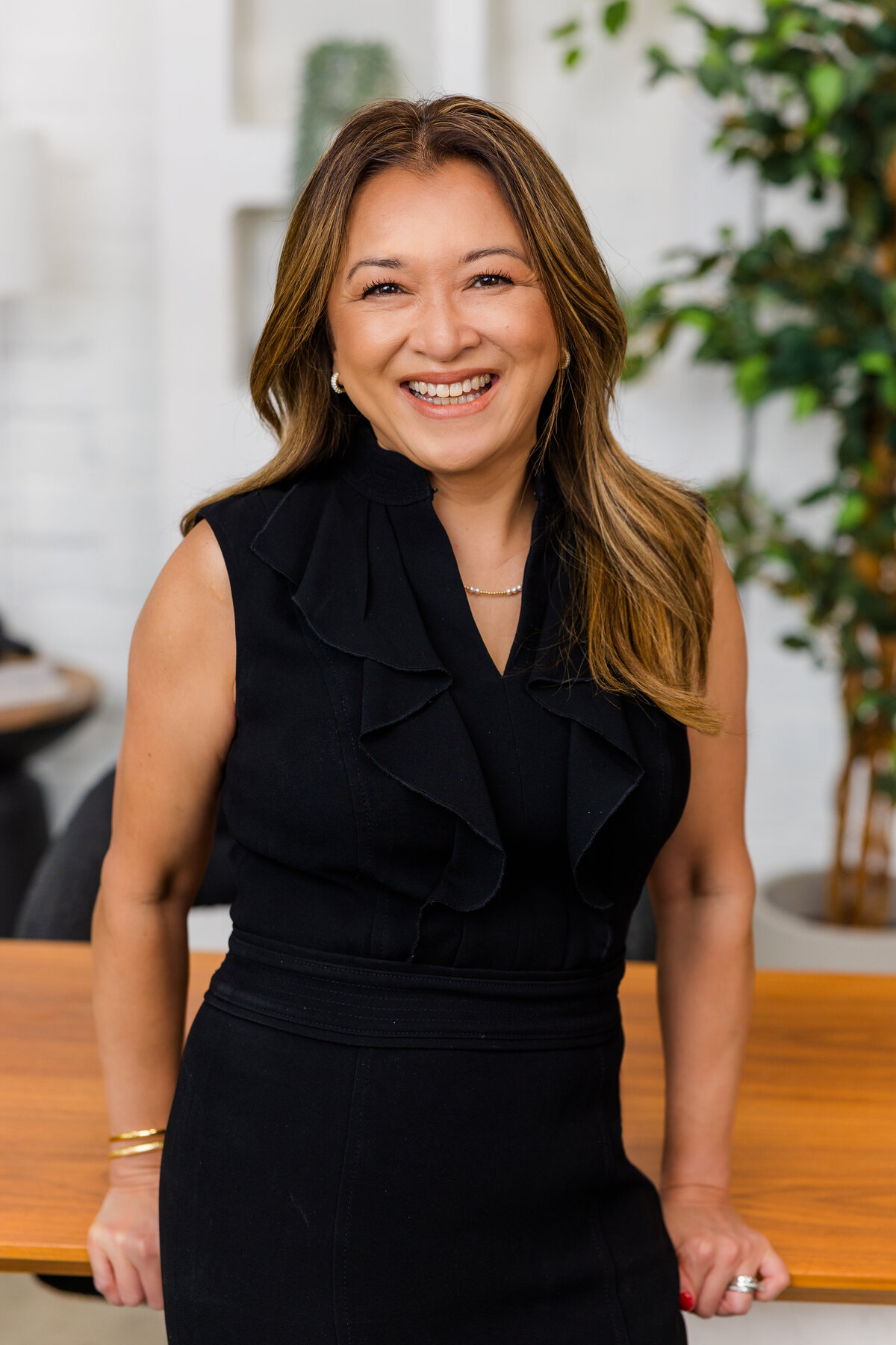 Corporate portrait of woman wearing a black dress leaning against an office desk with a lifestyle background