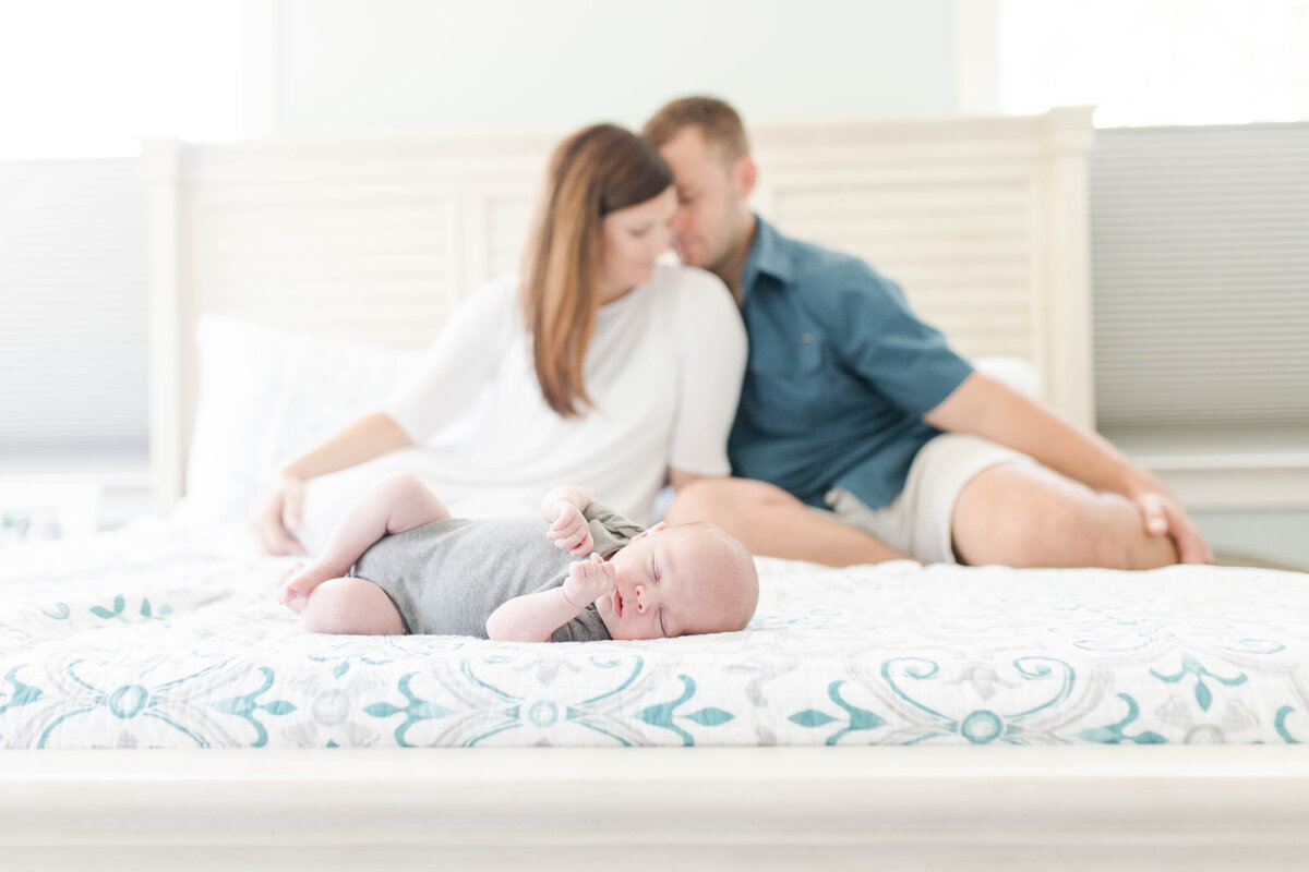 baby sitting on bed with parents behind photographed by Kaitlin Mendoza Photography, a family photographer in Carmel, Indiana