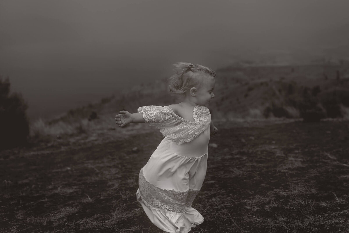 A little girl wearing a long white dress stands with her arms spread wide enjoying the wind. Captured in black and white by Brittany Danielle