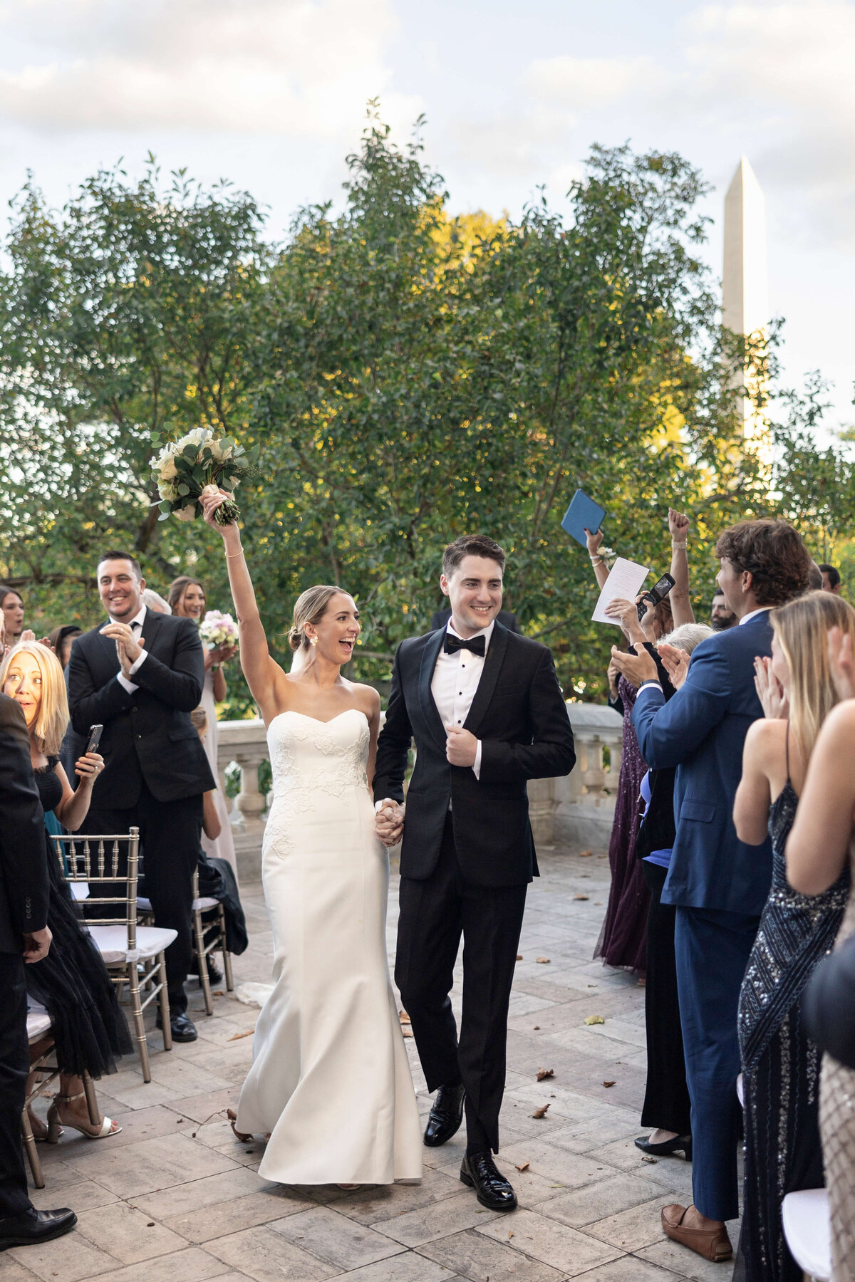 A newlywed couple celebrates as they walk down the aisle outdoors. The bride holds a bouquet aloft, wearing a strapless white gown, and the groom is in a black tuxedo. Guests on both sides cheer and clap. Trees and a monument are visible in the background.