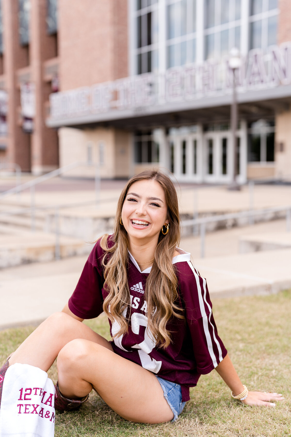 Texas A&M senior girl laughing while sitting on ground in front of Kyle Field and wearing Aggie maroon jersey and 12th man towel