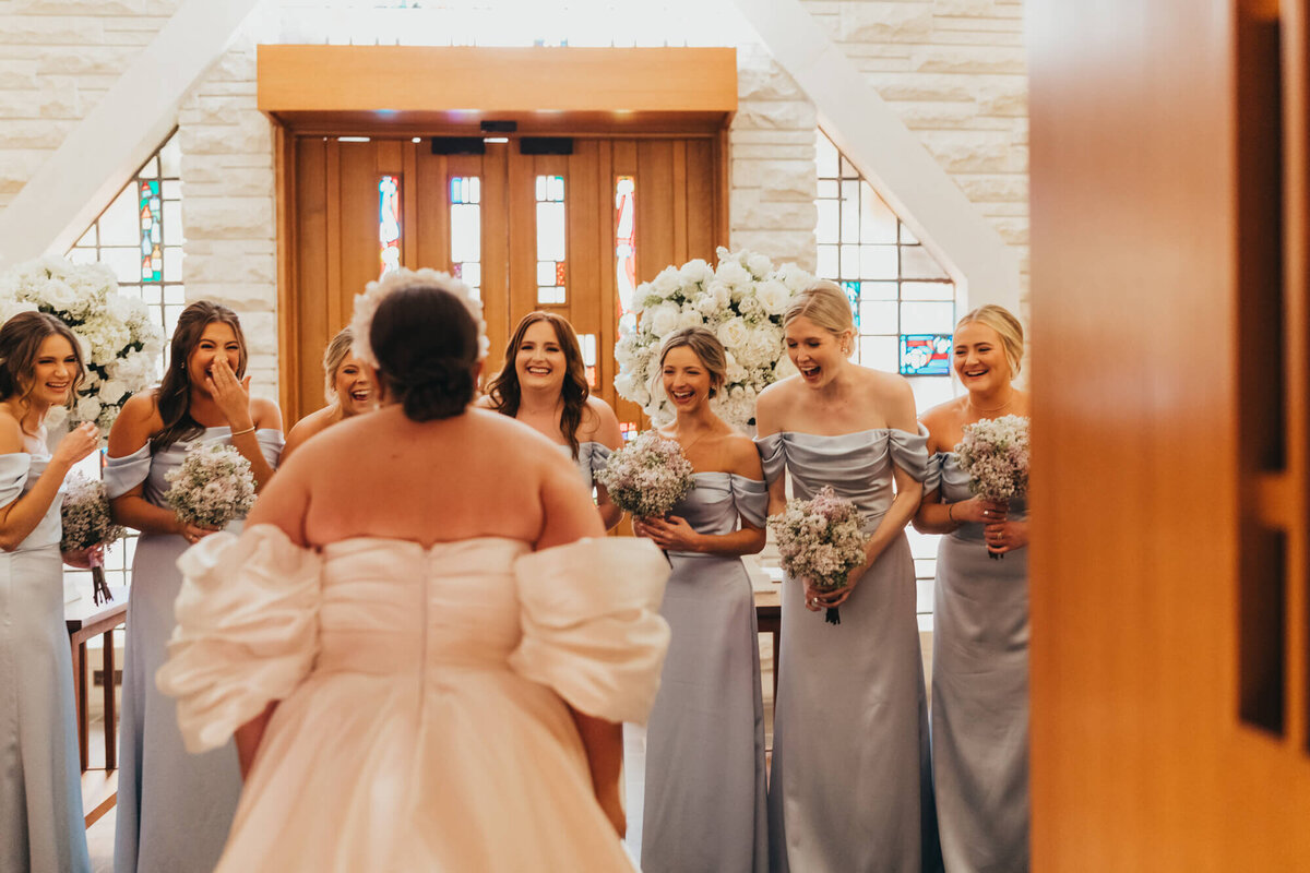 bride reveals her dress to her bridesmaids as they show their excitement in church.
