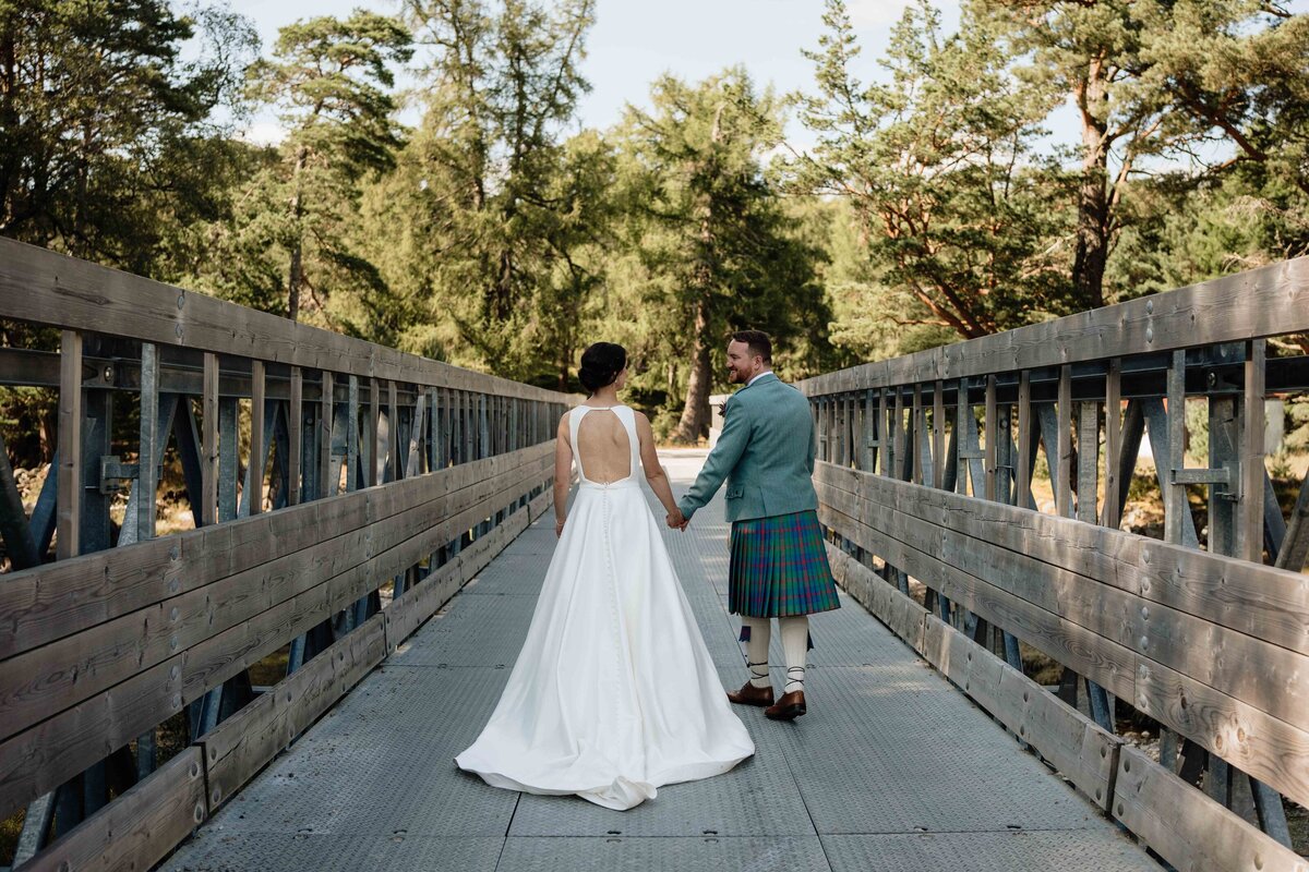 A wide photograph of a couple walking hand in hand across a wooden bridge at Mar Lodge.