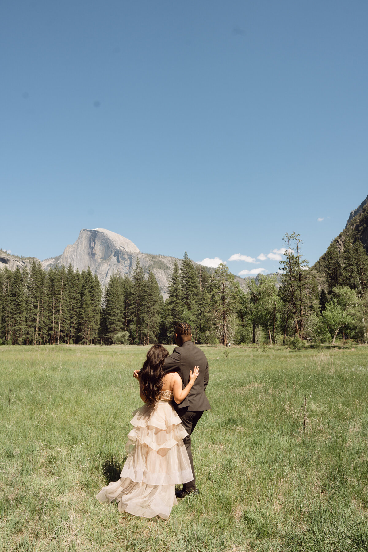 Engaged couple in meadows of Yosemite looking towards the silhouettes of the mountains with her tiered pink tulle gown on