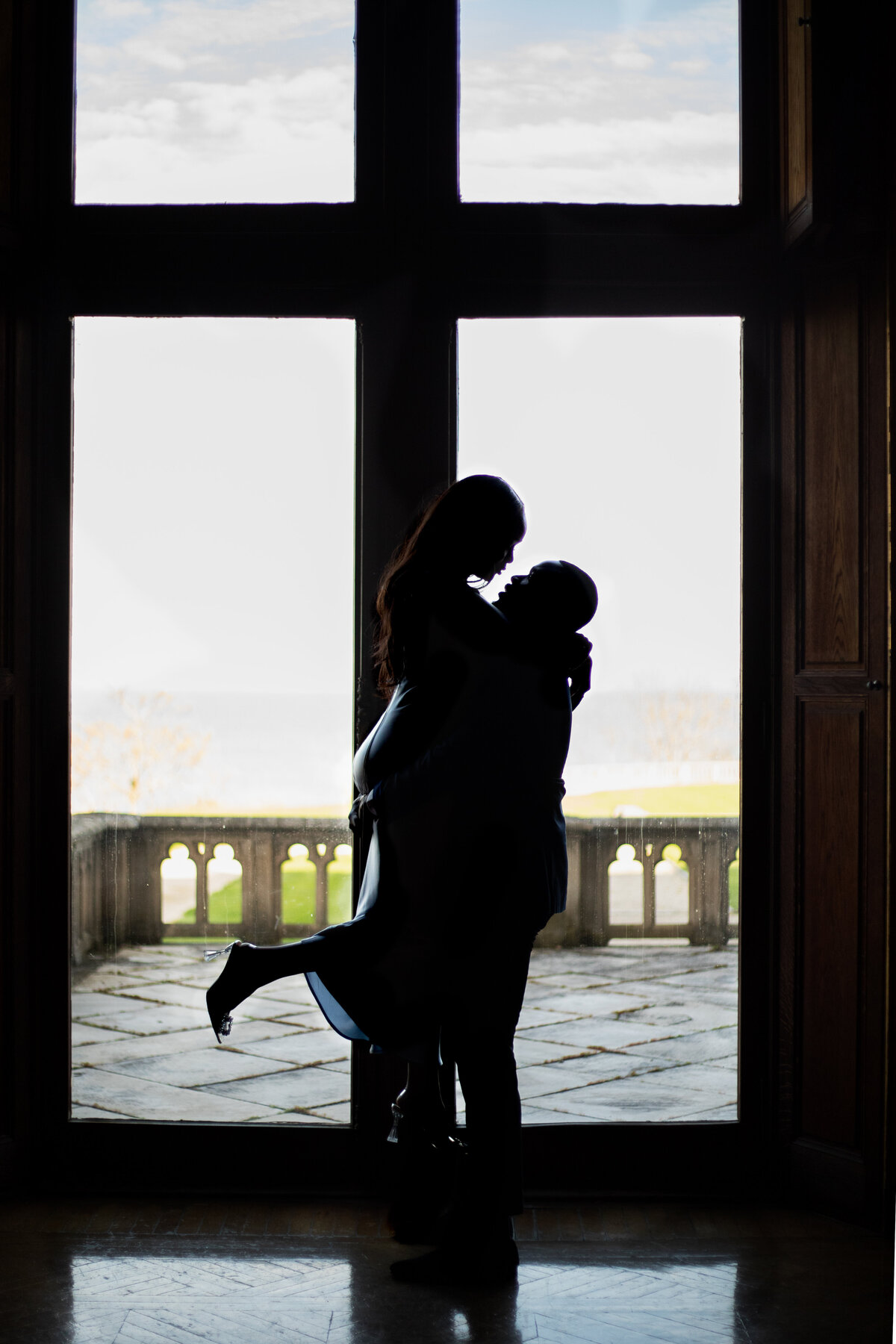 Ochre Court Mansion  | Kelsey Sheehan Photography Timeless Rhode Island Weddings | A silhouetted couple embraces joyfully in a large doorway, with a bright outdoor landscape of grass and trees in the background. Romantic and serene.
