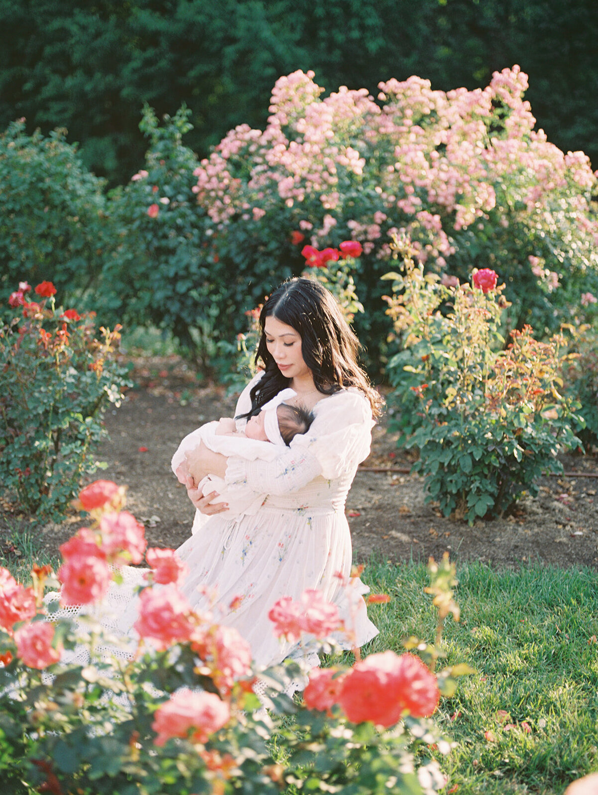 Dark-haired mother kneels in a rose garden as she holds her baby girl.
