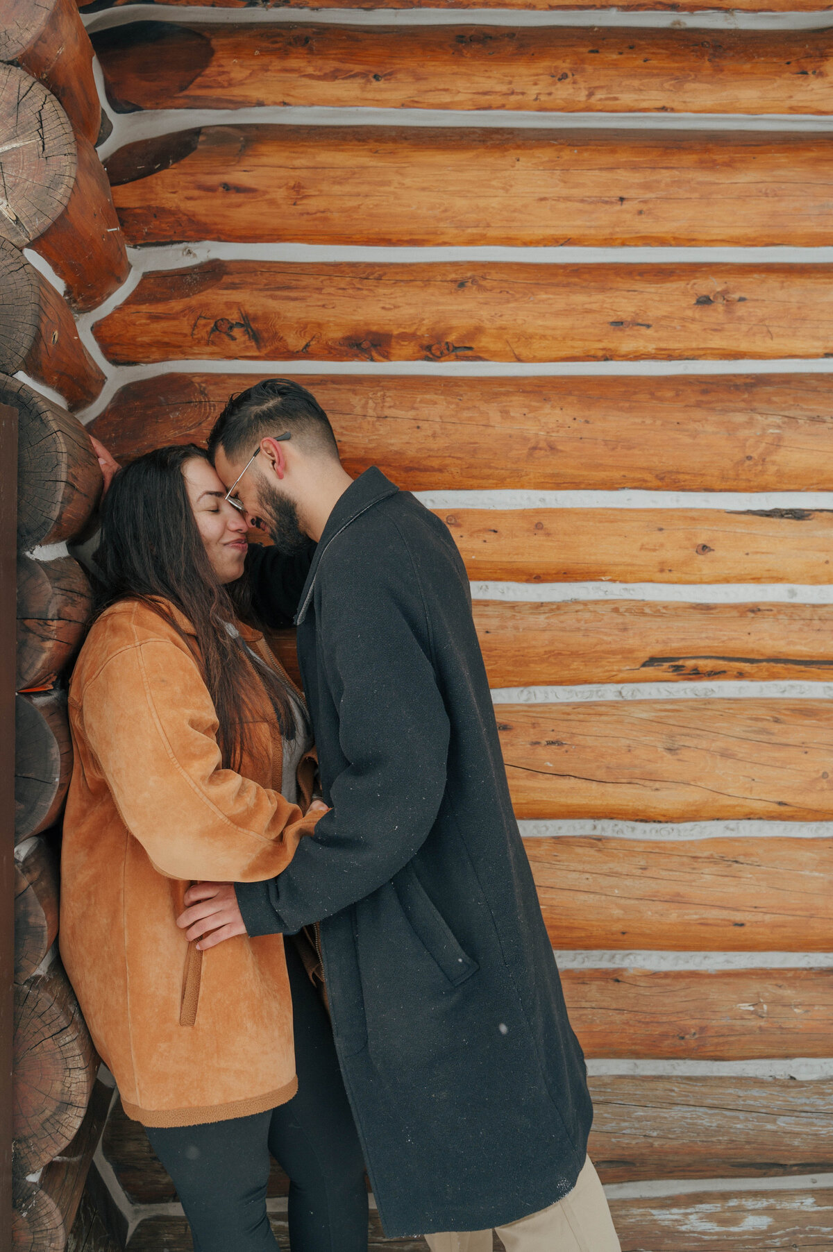Engaged couple leans in close beside a rustic log cabin at Lake Louise.