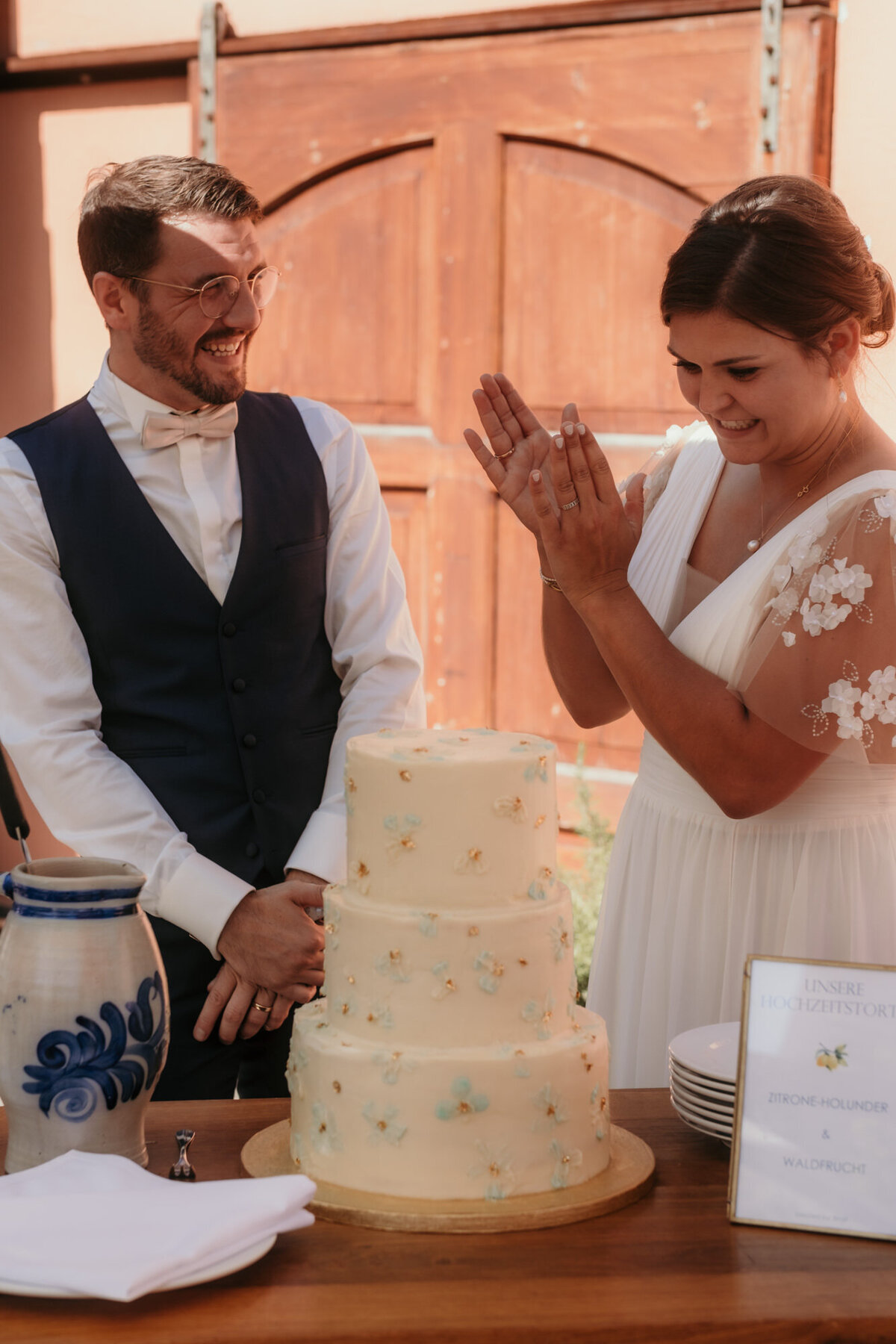 Das Hochzeitspaar steht vor ihrer Torte. Die Braut klatscht freudig in die Hände.