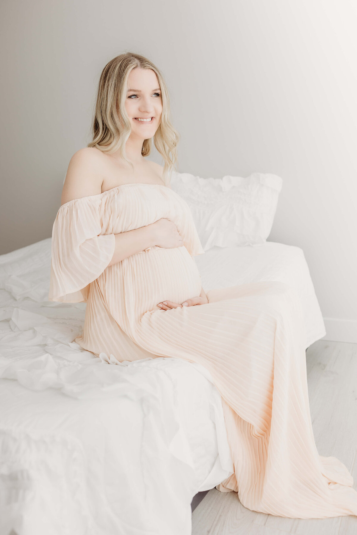 Blonde mom in light pink gown sitting on bed and smiling