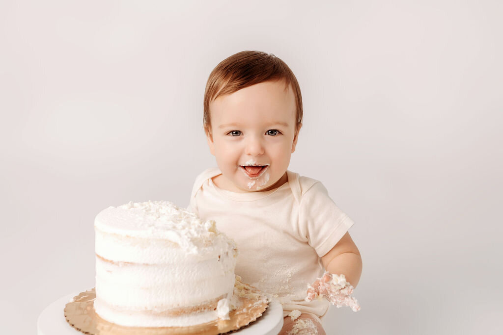 Baby boy with cake covered smile sits behind partially destroyed white frosted first birthday cake during timeless portrait session