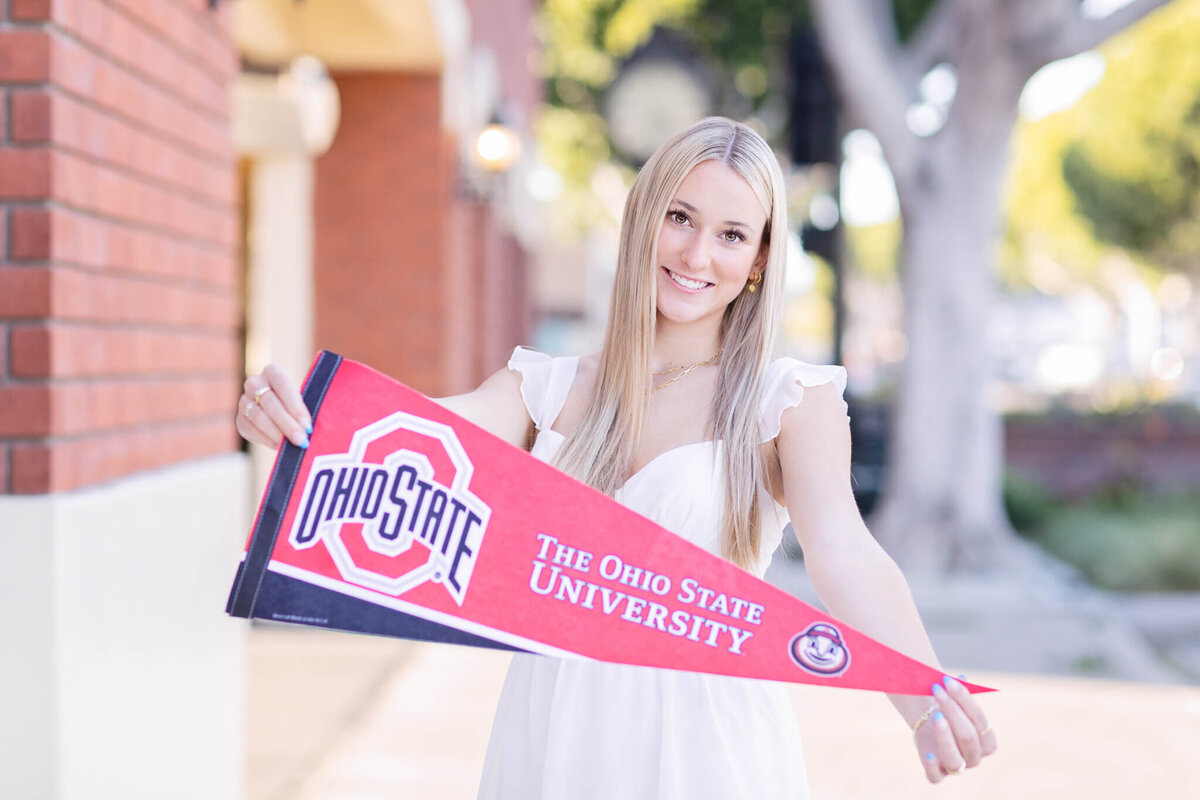 A person smiling and holding an Ohio State University pennant. They are outdoors, with brick walls and trees in the background.