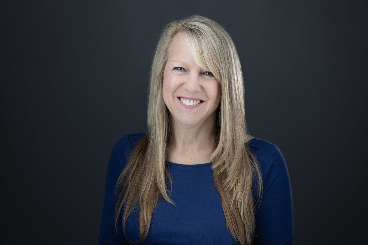 A headshot with a dark grey background of a woman with blond hair and smiling big at the camera. She is wearing a blue shirt and projecting confidence and approachability. Photo by sacramento headshot photographer, philippe studio pro.