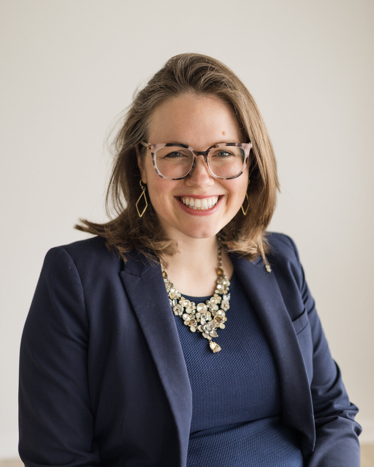 Brunette blonde pregnant female sitting in a chair, wearing glasses, a dark blue dress with a long sleeve dark blue jacket, smiling at the camera