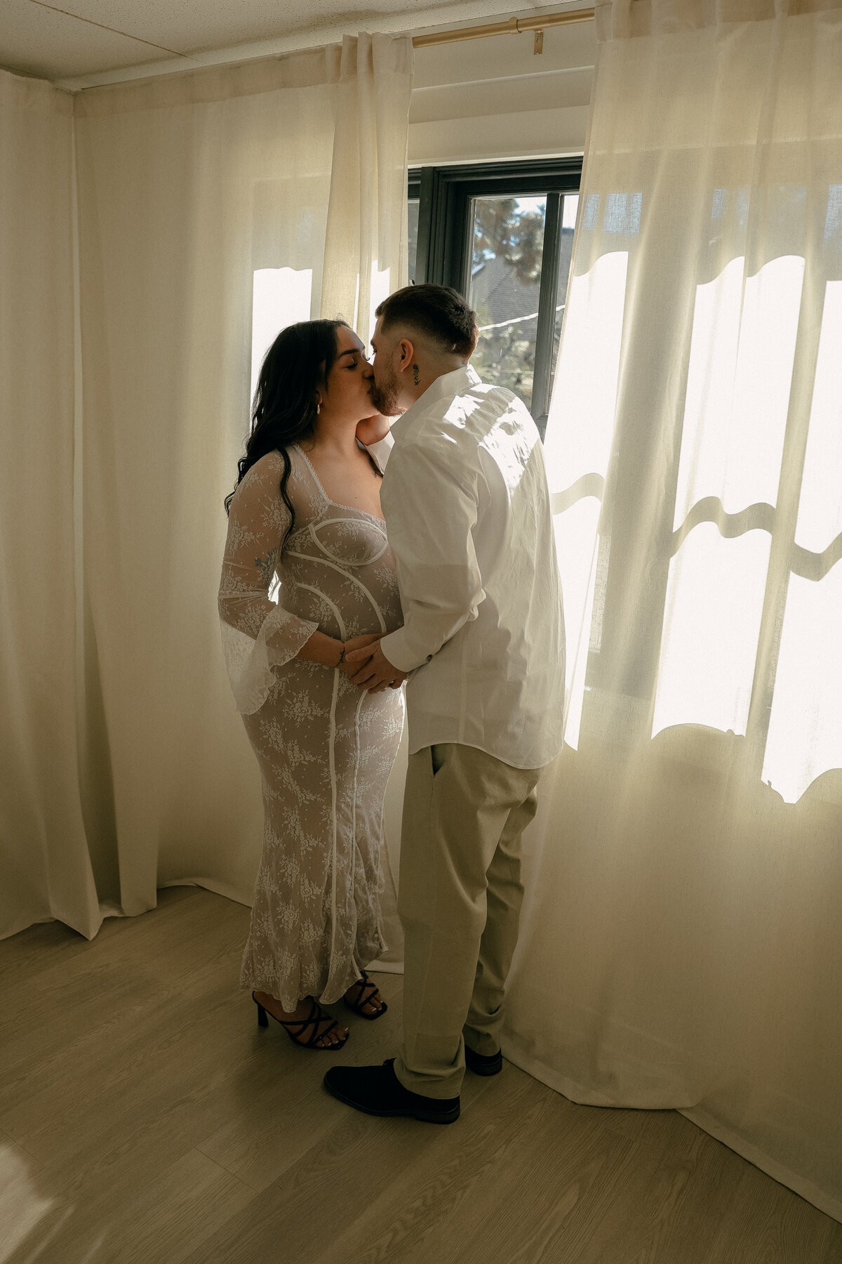 Indoor Studio Maternity Session with Couple Kissing Beside Sheer Curtains and Warm Sunlight