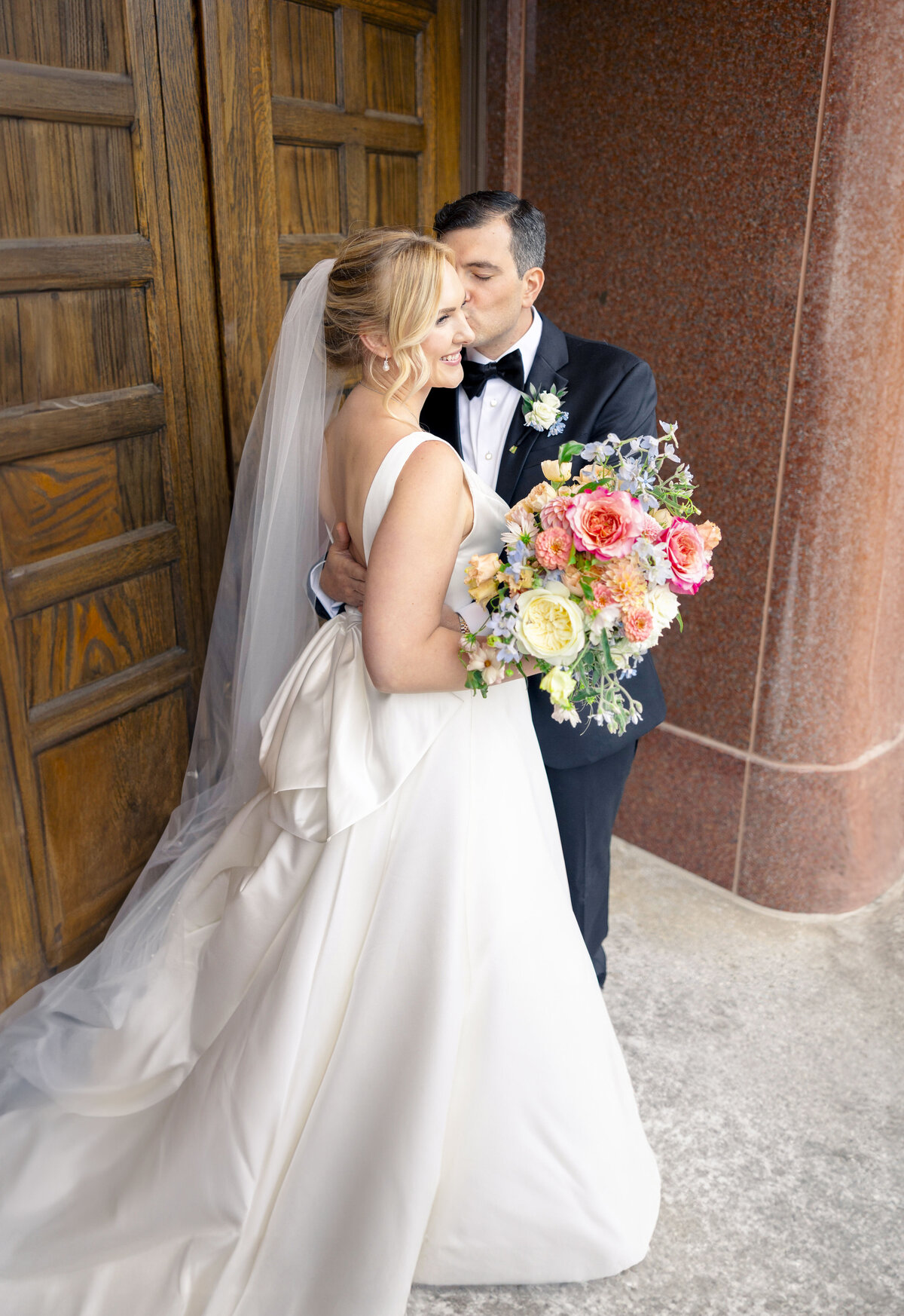 A bride and groom embrace outside a building. The bride wears a white gown and veil, holding a bright bouquet with pink, orange, and yellow flowers. The groom is in a dark suit and kisses her on the temple. Wooden doors and stone wall are in the background.