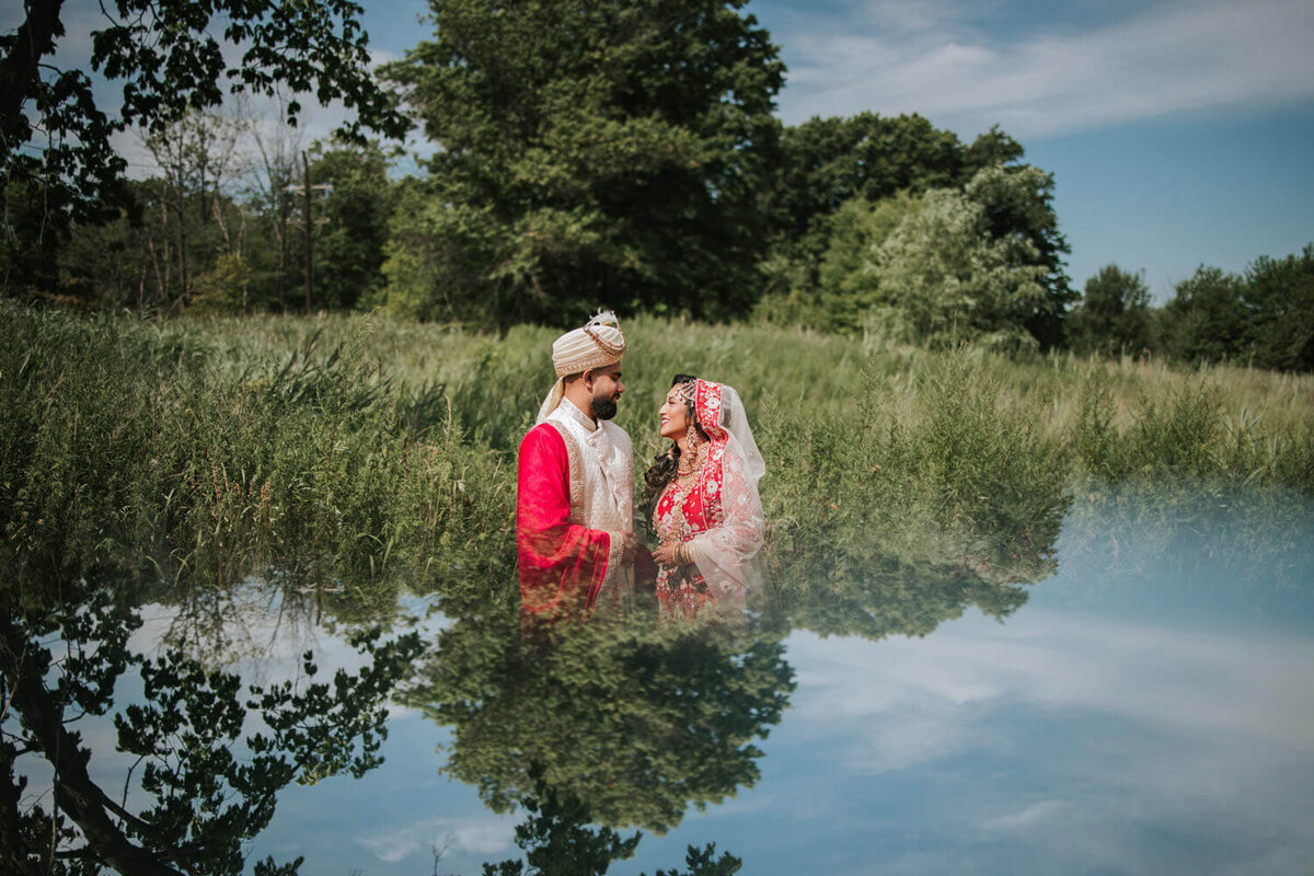 A couple dressed in traditional attire shares a joyful moment in a lush green field with a serene reflection.