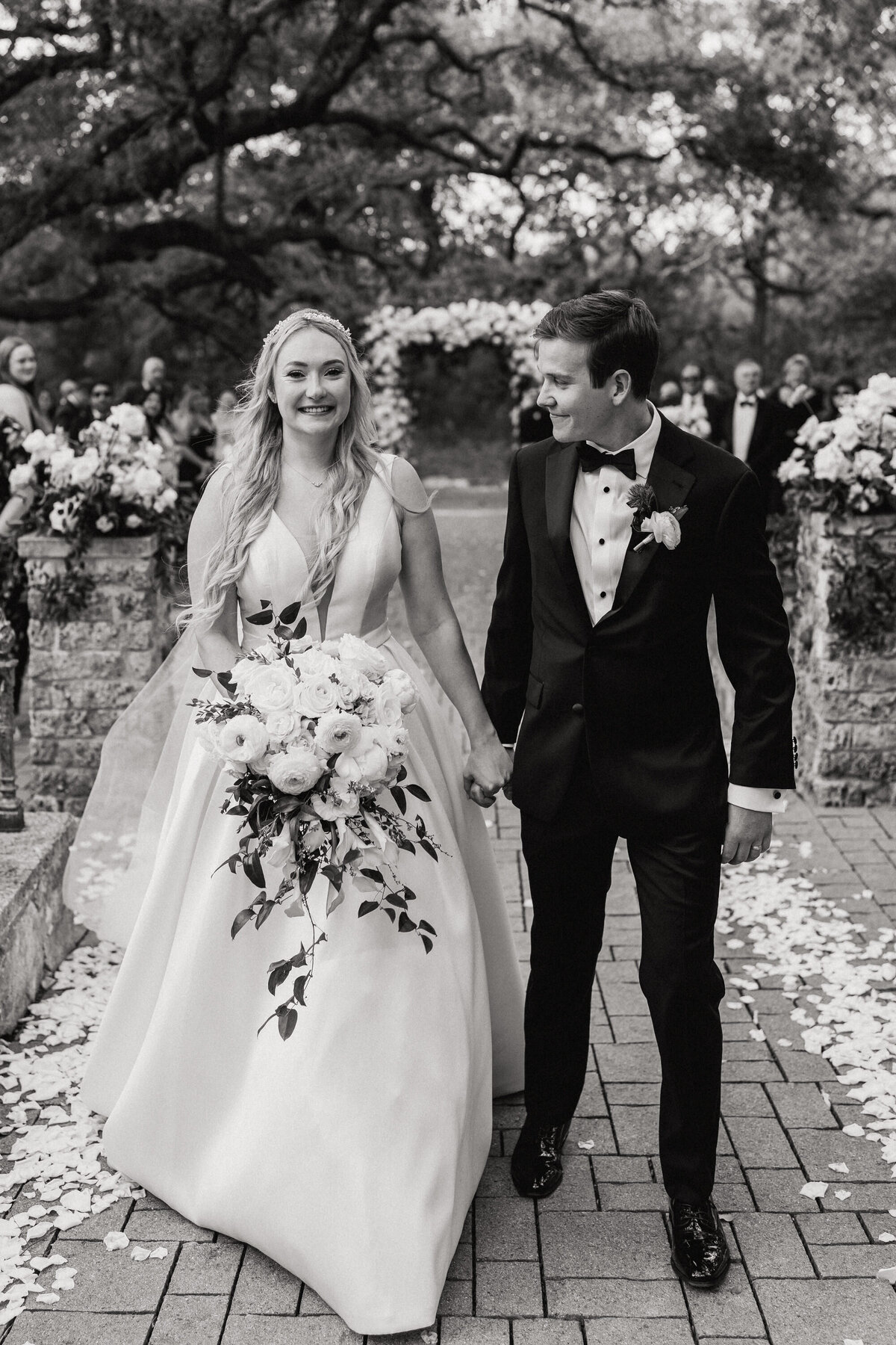 A black and white photograph of Maggie and Alex at Camp Lucy in Dripping Springs, near Austin, Texas. They bride and groom have just married and are holding hands as they walk through their guests down the aisle, which is lined with white rose petals. The bride is looking into the camera and smiling. She is wearing a satin gown, her light hair is long and wavy and she’s holding a white and green floral bouquet. The groom is smiling, looking at the bride and wearing a tuxedo. Wedding photography by Stacie McChesney/Vitae Weddings.