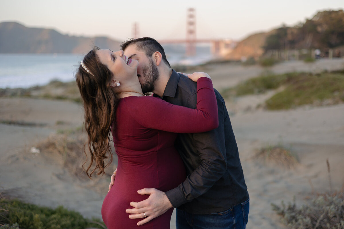 man hugging and kissing a womans neck in the field below the golden gate bridge