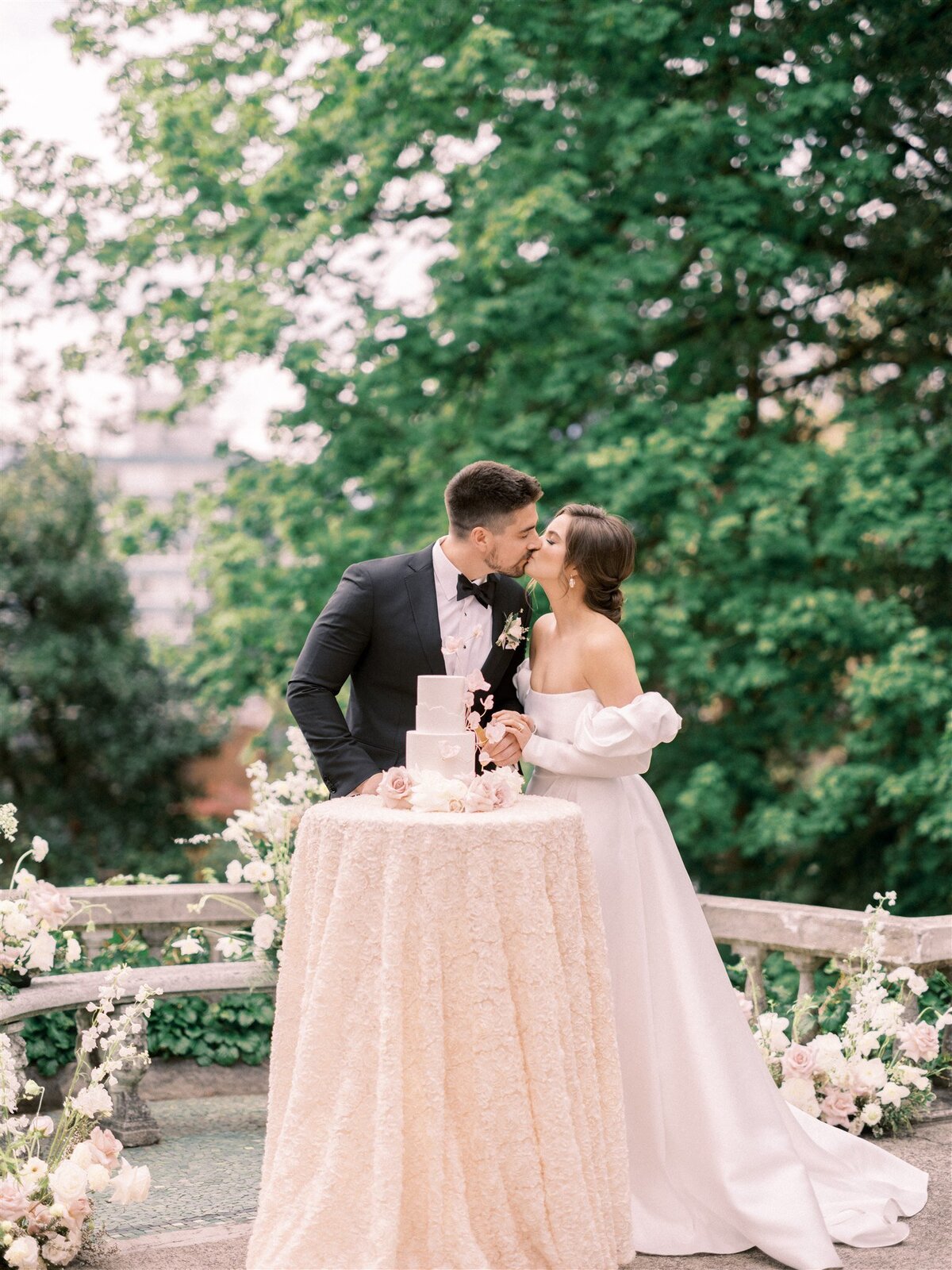 A bride and groom share a kiss beside a round table with a white cake. Organized by Destination Wedding Planner Melissa Dawn Event Designs, the couple is outdoors, surrounded by greenery and floral arrangements. The bride wears an off-shoulder white gown.