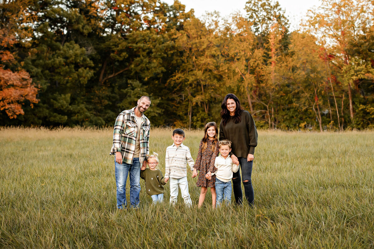 A family of six stands together in a grassy field. Behind them, autumn trees display orange and green foliage. The group includes two adults and four children, all casually dressed and smiling. The sky is clear, indicating a pleasant day.