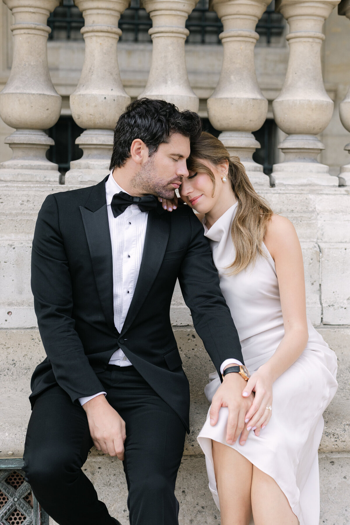 Couple at the Louvre in Paris during their engagement session, she rests on his shoulder.