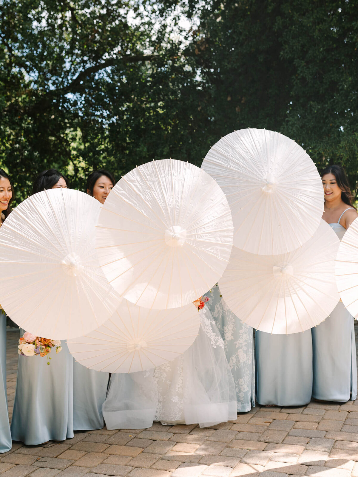 Bridesmaids in soft blue dresses hold white parasols, partially obscuring their faces. They stand on a brick path, framed by lush green trees.