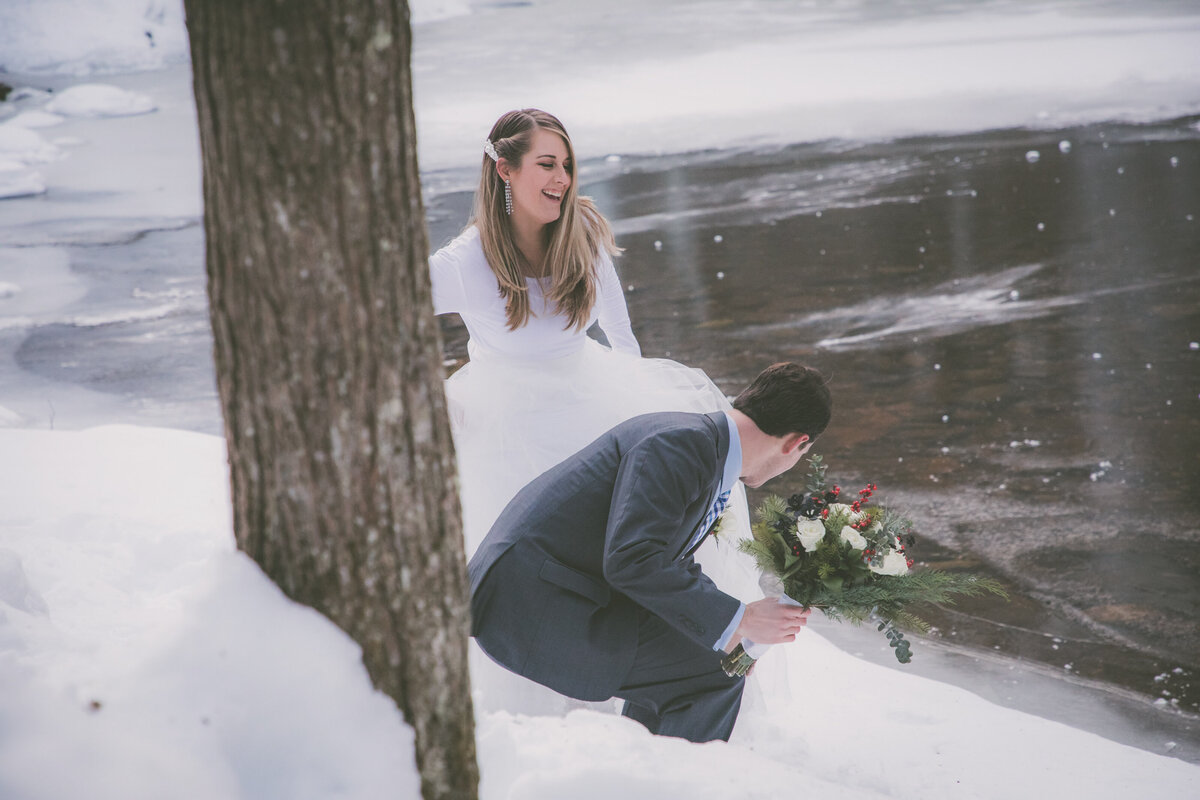 Groom assists bride while hiking through snow.