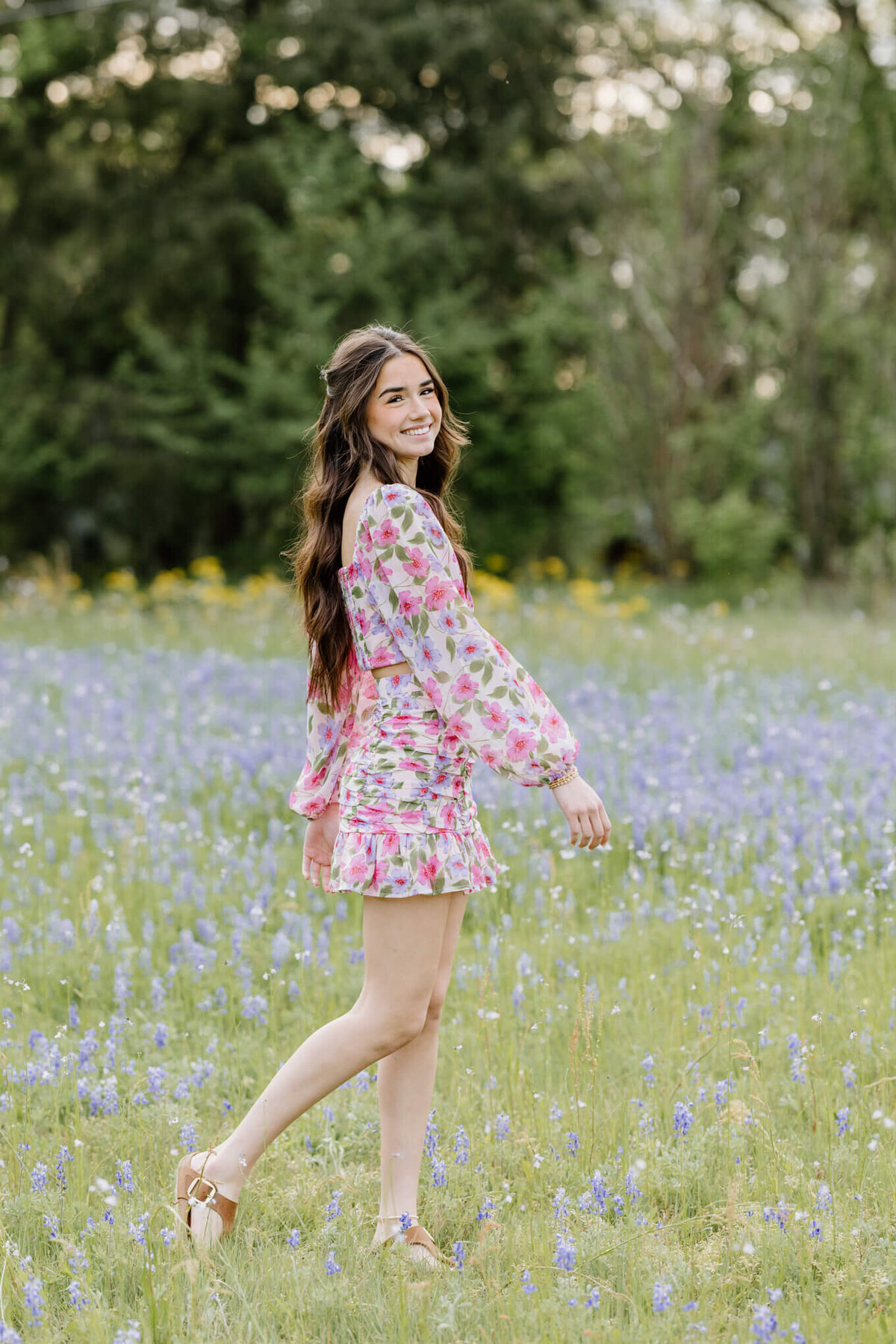 portrait of graduate in pink floral dress walking through field of bluebonnets