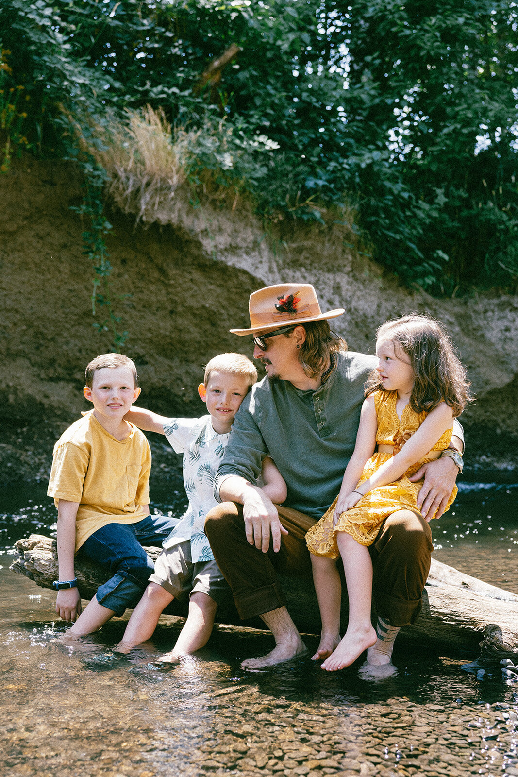 Family of four take casual candid summer photos sitting on a log in a river in Oregon.