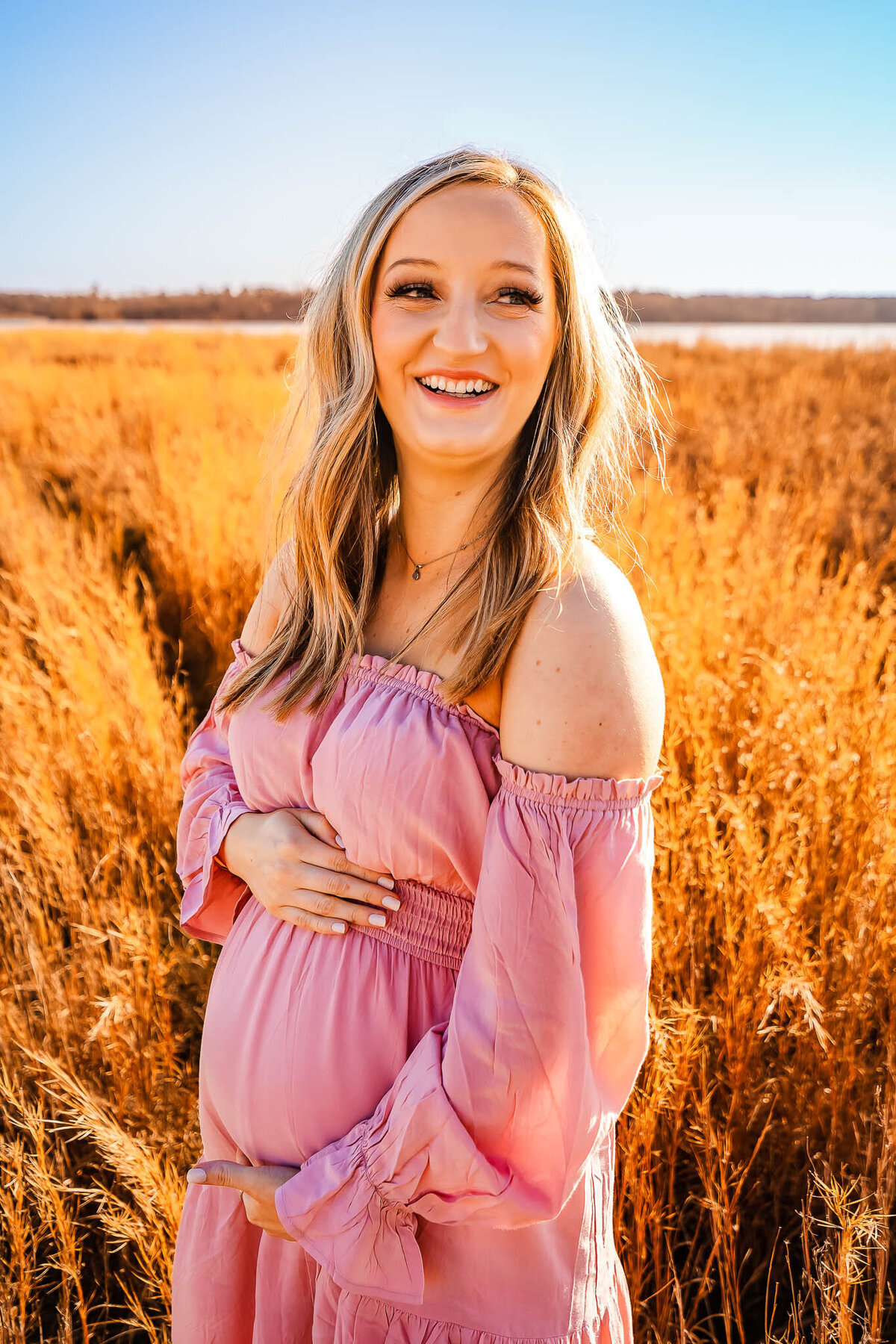 Laughing pregnant mother in a pink dress in a grassy field during golden hour