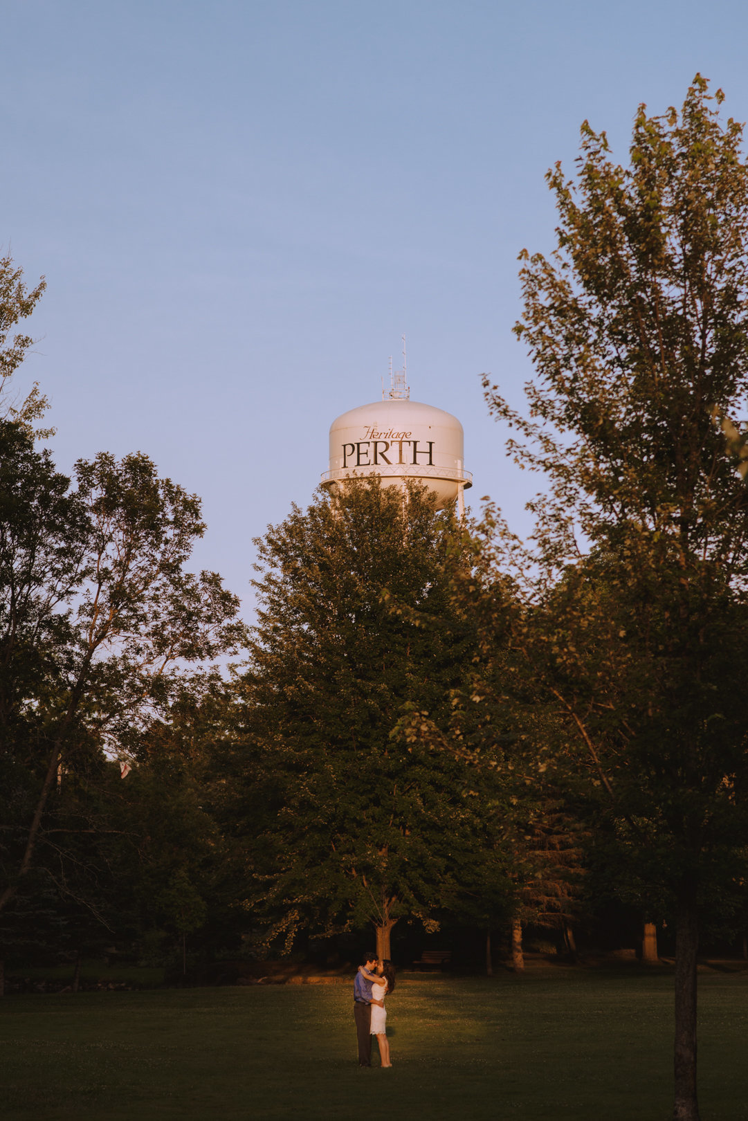 engaged couple standing in a path of sunlight in Stewart's Park with Perth water tower behind them