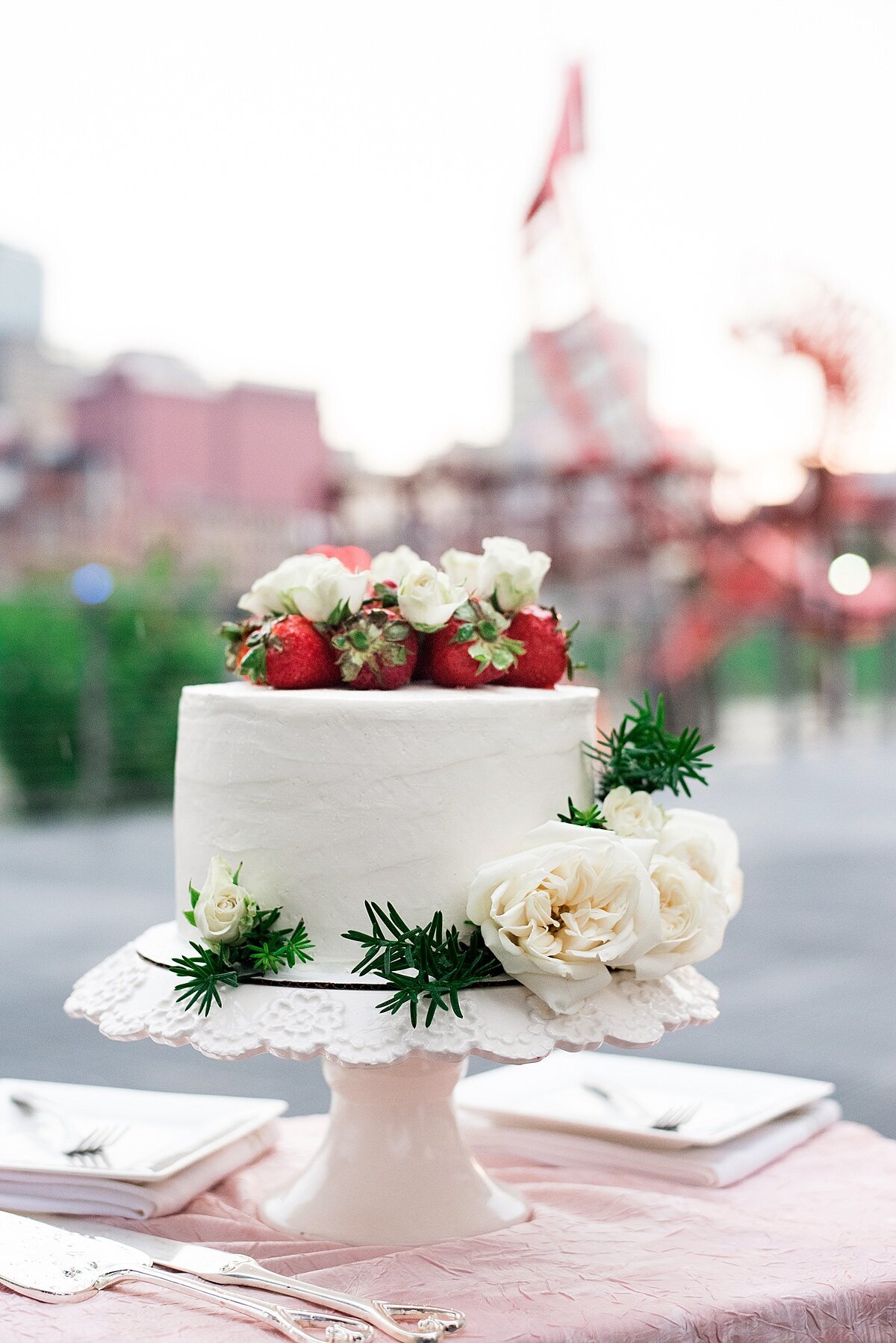A one tier white wedding cake sits on a white pedestal cake stand. The cake stand is placed on a table with a pink table cloth, two white square plates with forks and a silver cake knife and server set. The cake is decorated with rosemary, white roses and strawberries.