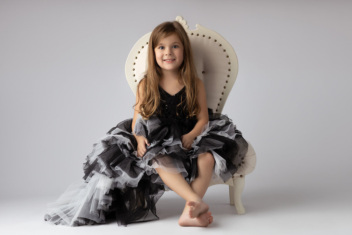 A smiling young girl in a black and gray ruffled dress, sitting on an elegant chair with bare feet.