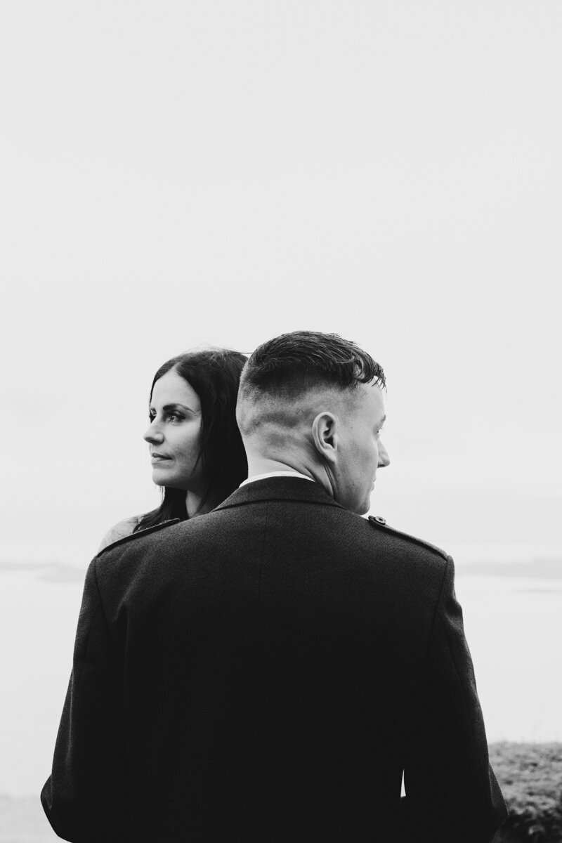 Bride and Groom standing on a mountain top. The wind is blowing in her hair and they are looking out to the view while hugging each other. The bride is a long sleeved wedding gown.