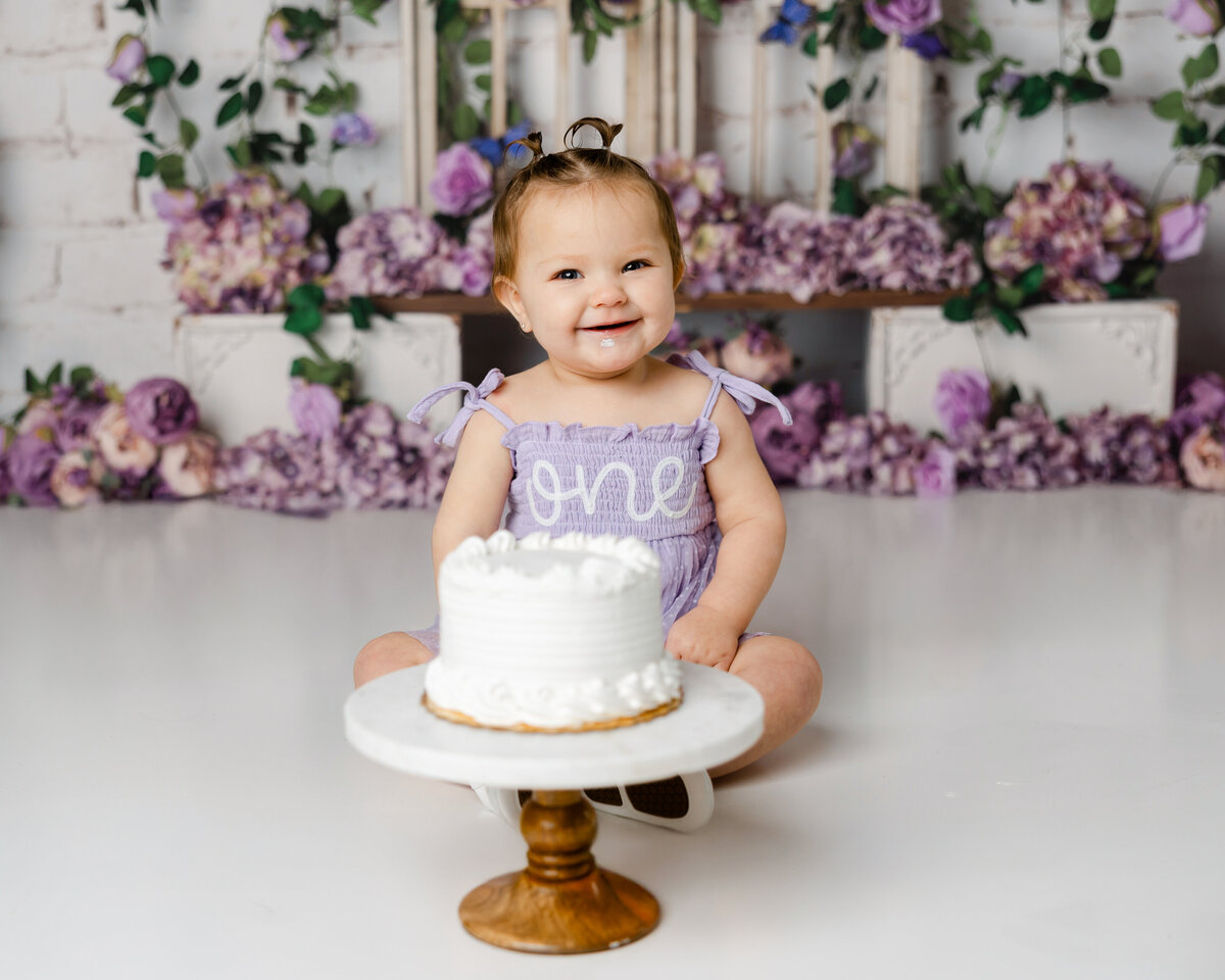 Smiling baby in lavender “one” dress with a white cake and purple floral backdrop. Photograph by Yucaipa cake smash photographer Kaitlyn Dawn Photography.