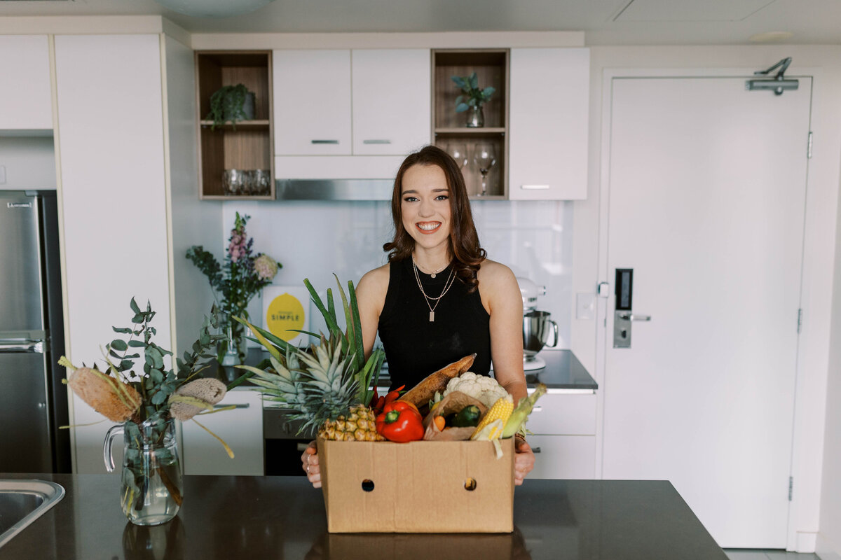 Dietitian Annabel holding a box brimming with fresh produce, promoting whole food nutrition and wellness.