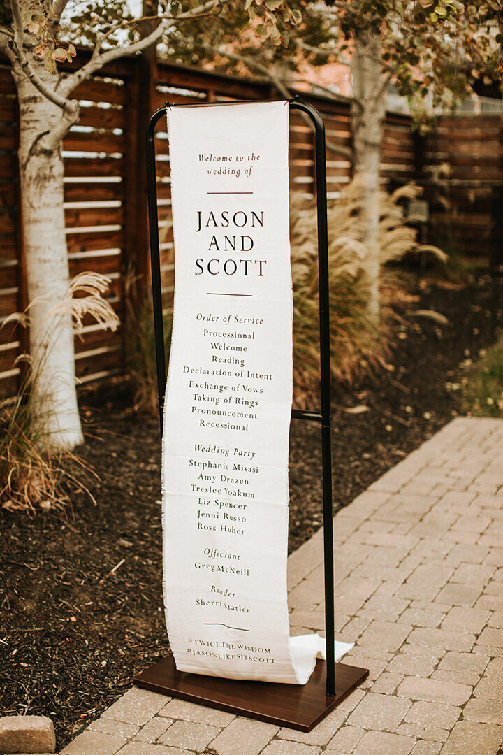 White wedding banner with black font hanging on black metal stands in the outdoors.