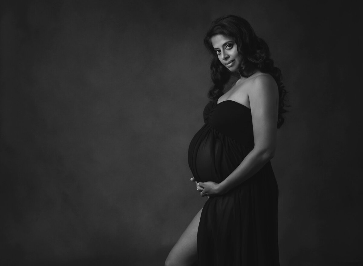 maternity photo of woman wearing black dress holding her belly on dark background
