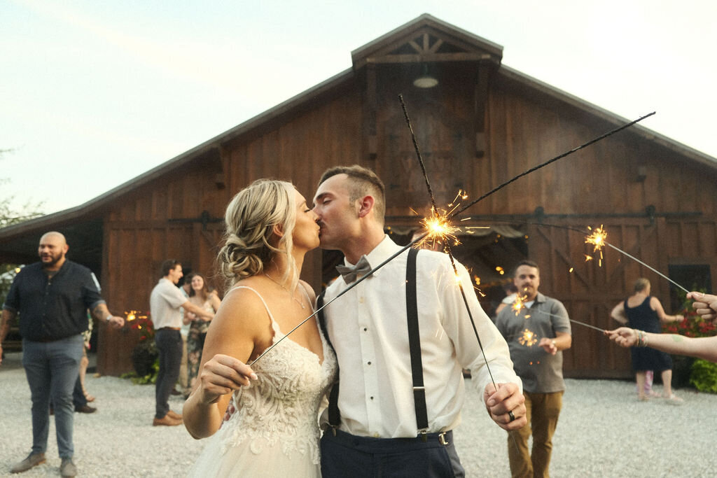 Bride and groom sharing a sparkler kiss in front of a barn with guests
