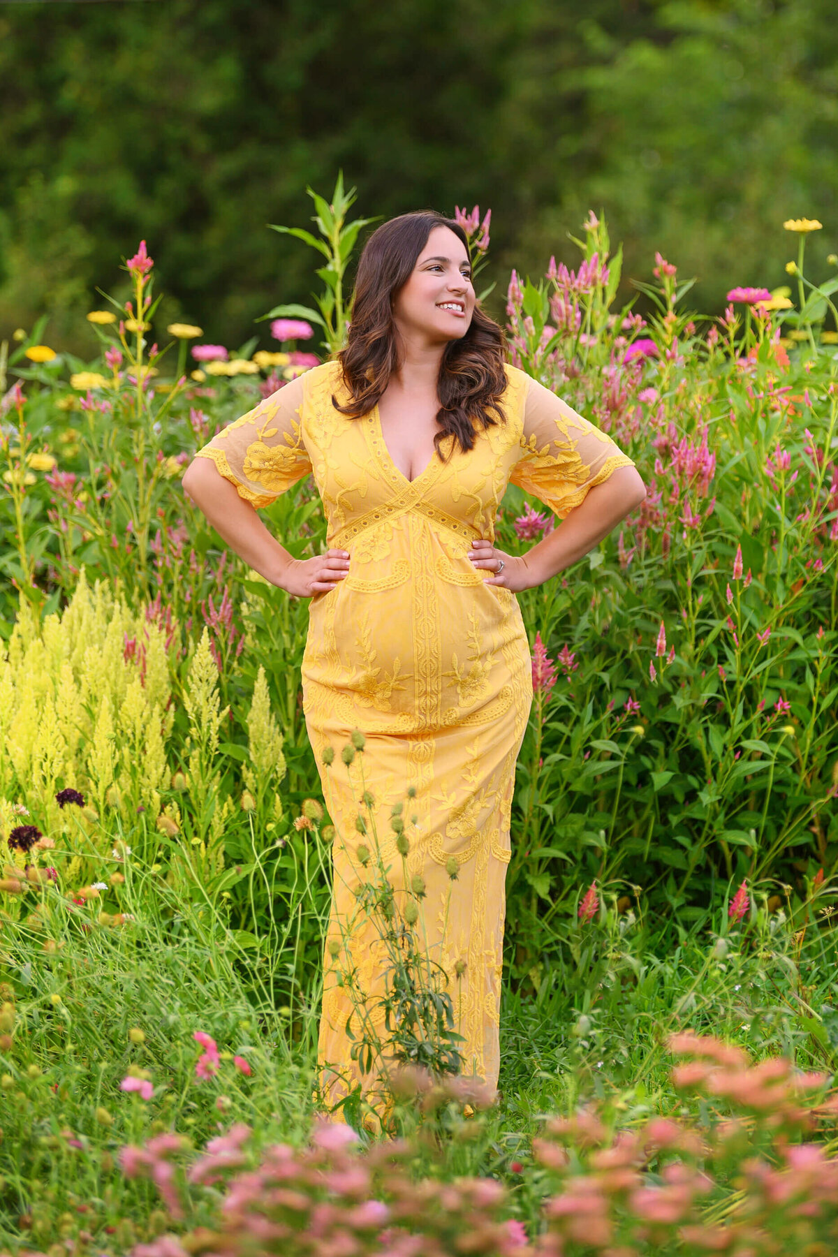 Brown-haired pregnant woman wearing a yellow dress with her hands on her hips smiling for her outdoor maternity photography session in Burlington, Ontario