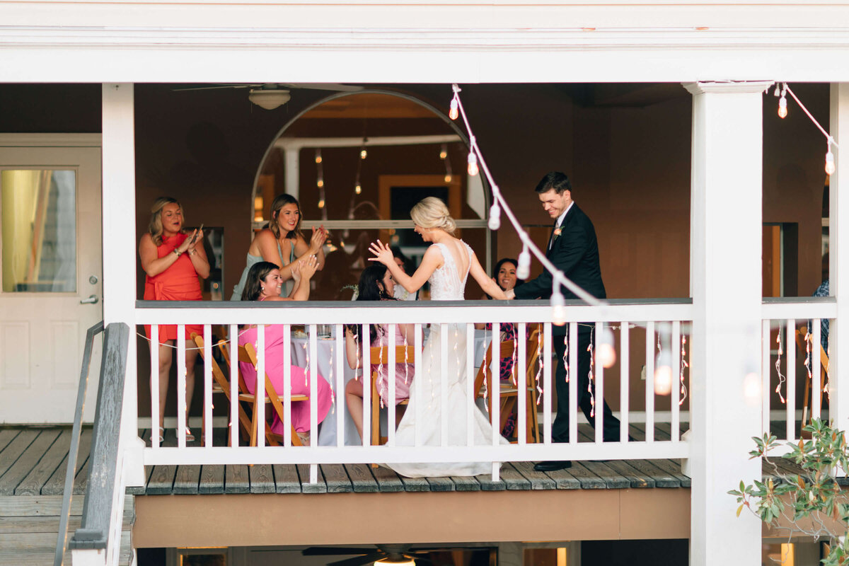A bride and groom walk onto a balcony where guests seated at tables clap and cheer for them. The scene is festive, with string lights and the couple in wedding attire, creating a joyful celebration atmosphere.