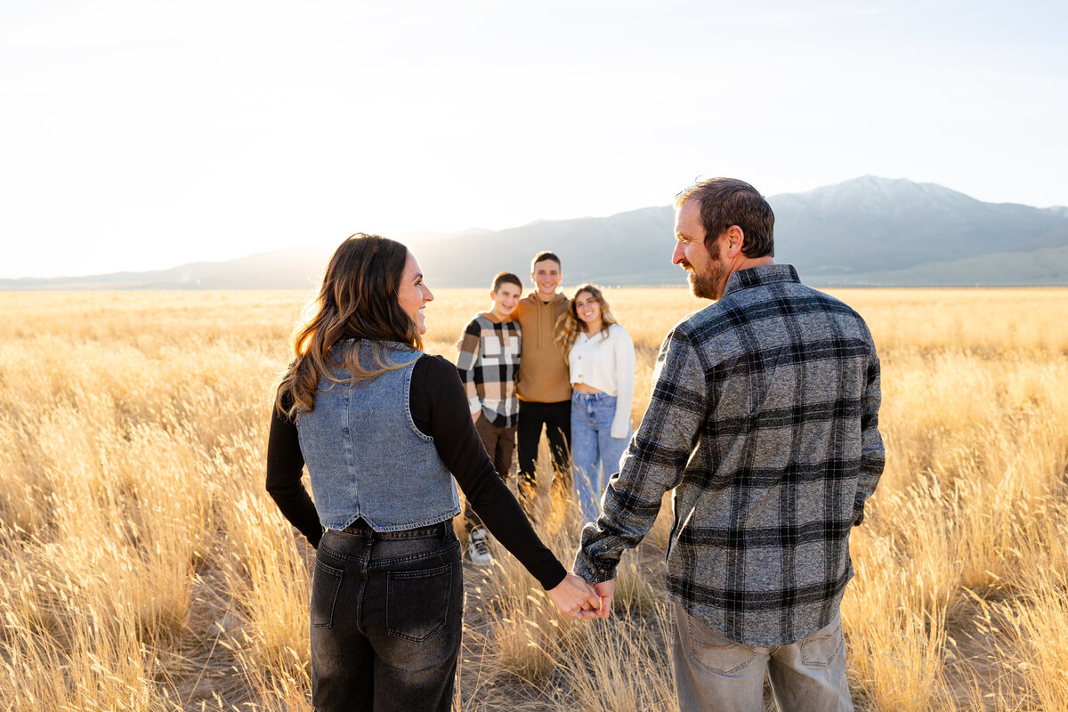  A couple holds hands in a sunlit field in Eagle Mountain, Utah, looking back at their three children standing in the distance with mountains in the background.