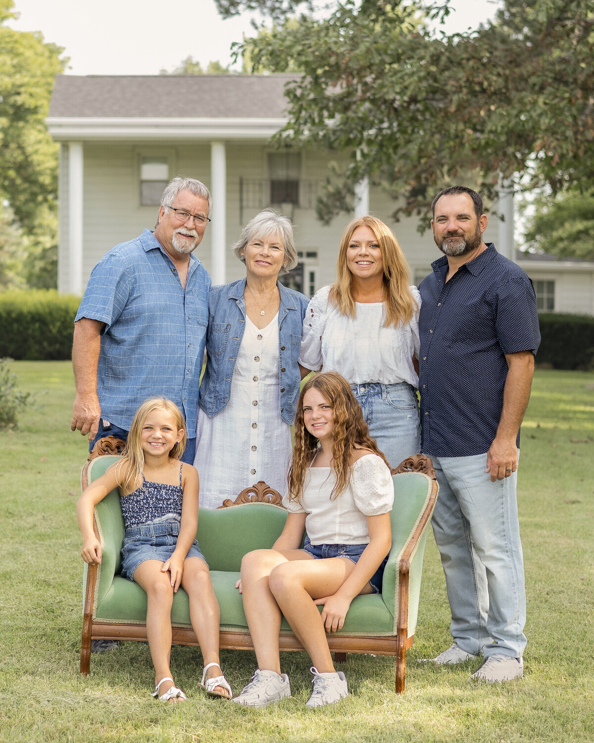 Grandparents, mom, and dad are standing behind the vintage green couch in the grass with their family home in the background.  Daughters are sitting on the vintage green couch.  Everyone is looking at the camera smiling.  Mom is wearing a jean skirt, white half sleeve shirt, and has long blonde hair.  Dad is wearing a blue button up short sleeve shirt, blue jeans, and has black gray short hair and beard.  Older daughter is wearing blue jean shorts, a white short sleeve shirt, and has long brown hair.  Grandpa is wearing a light blue and white stripped short sleeve button up shirt, blue jean shorts, and has white short hair and goatee.  Grandma is wearing a white button up dress with a jean jacket over it, with short white hair.  Youngest daughter has a dark blue and white tank top style shirt with a blue jean skirt, and has medium length blonde hair.